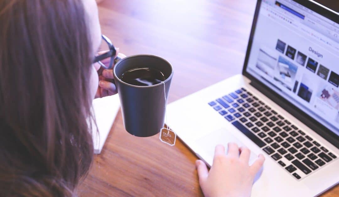 A woman enjoys coffee while browsing the web on a laptop at a home office desk.