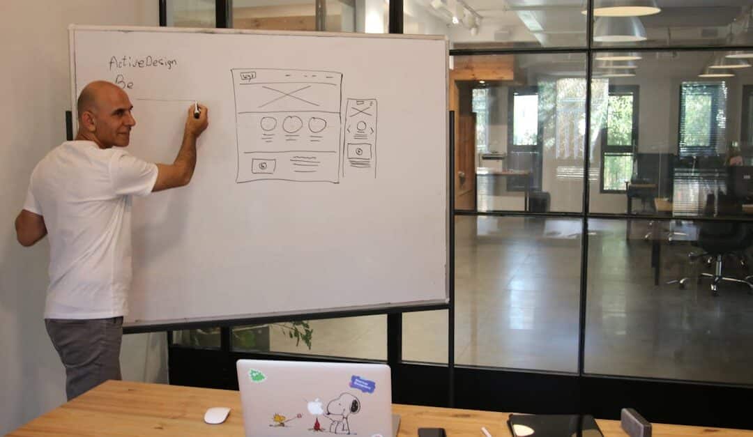 Side view of middle aged male leader in white t shirt writing on whiteboard during presentation in modern office space with glass walls
