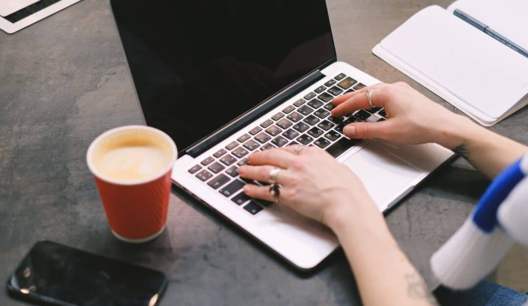 A freelancer typing on a laptop with a cup of coffee, smartphone, and notepad on the table.