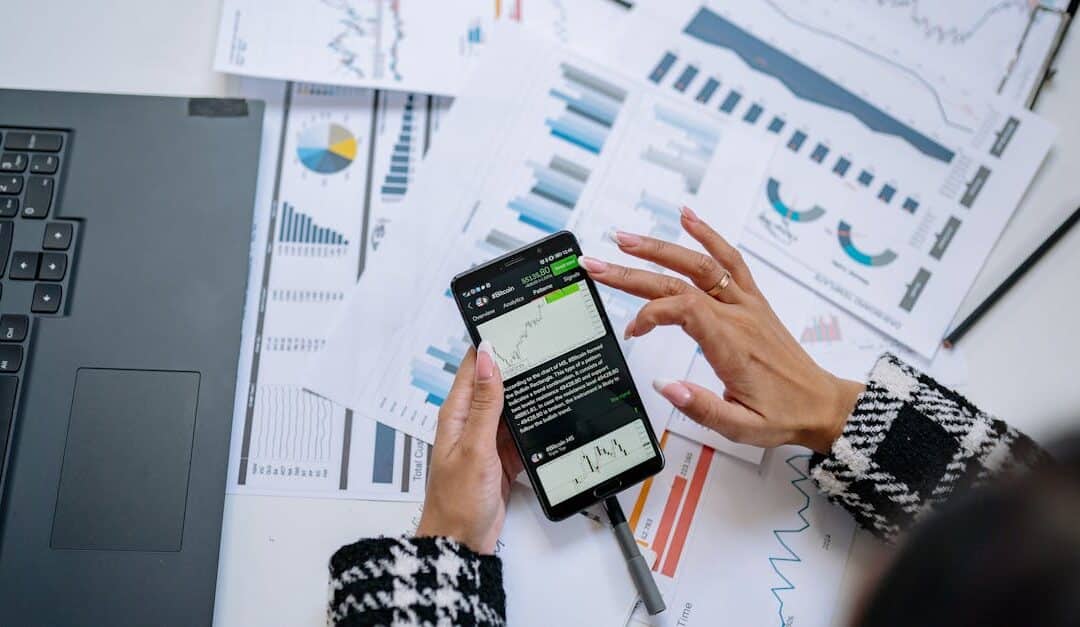 Businesswoman analyzing financial charts on smartphone with laptop and documents.