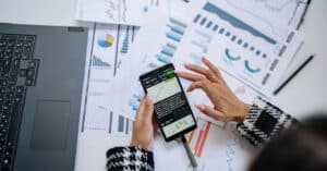 Businesswoman analyzing financial charts on smartphone with laptop and documents.