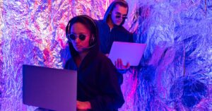 Two individuals wearing hoodies working on laptops in a neon-lit, futuristic room.