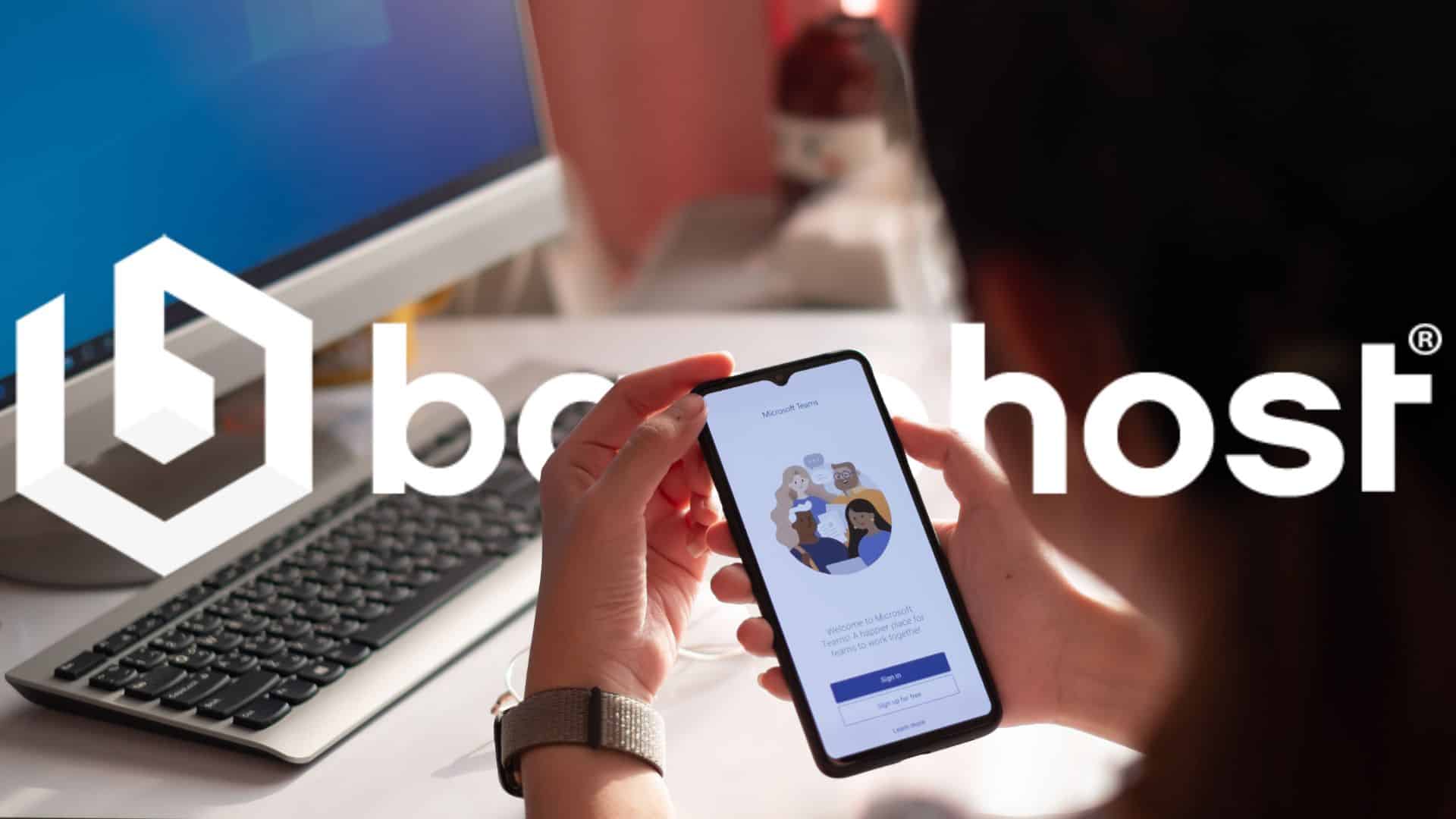 Person holding a smartphone with Microsoft Teams open, sitting at a desk with a computer, featuring the BaseHost logo overlay.