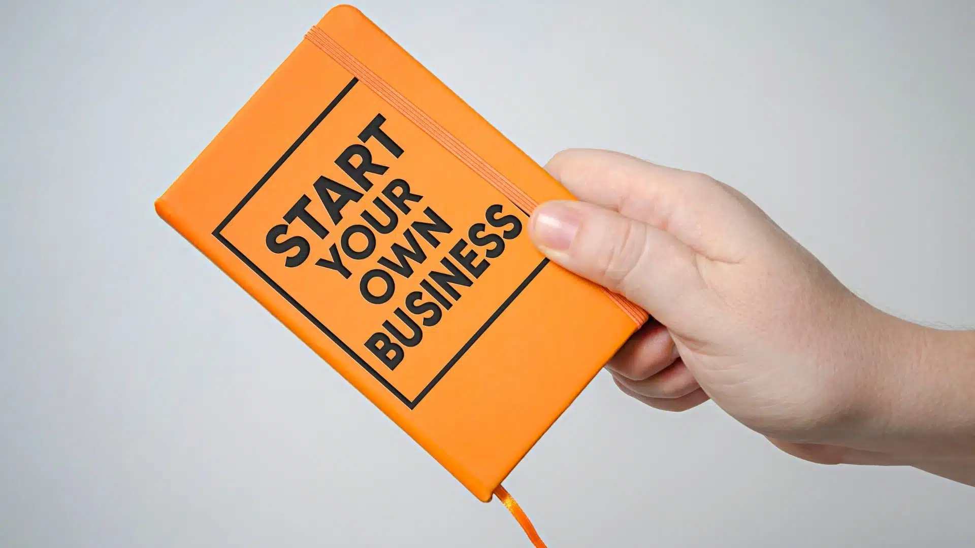 hand holding an orange notebook with the words start your own business, representing starting a business in 2026